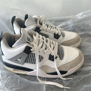 Nike Air Jordan 4 vit/svart/grå - Säljer ett par Nike Air Jordan 4 sneakers i vitt, svart och grått. Skorna har klassisk meshpanel, snörning och detaljer i mocka och skinn. Snygg retrostil med AIR-logga bak och coola plastdetaljer på sidorna. Perfekta för dig som gillar streetwear och ikoniska sneakers.