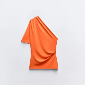 Orange oneshoulder topp från Zara - Säljer en snygg orange topp från Zara med oneshoulder-design. Toppen har en kort ärm på ena sidan och är ärmlös på andra, vilket ger en cool asymmetrisk look. Tillverkad i mjukt och stretchigt syntetmaterial som sitter skönt mot kroppen.