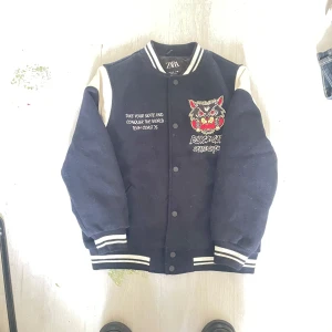 Svart bomberjacka från Zara med brodyr - Cool svart bomberjacka från Zara med vita detaljer och broderad 'Black Cats Skate Crew' på bröstet och ryggen. Jackan har tryckknappar, ribbade muddar och kontrastfärgade ärmar. Perfekt för dig som gillar streetstyle och skatekänsla.