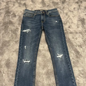 Jack & Jones jeans  - Snygga blå jeans från Jack & Jones i modellen Skinny Liam men sitter mer slim . Jeansen har slim passform, slitna detaljer och klassisk femficksdesign. Materialet är stretchigt denim med orangea kontrastsömmar och medelblå tvätt. Perfekta för dig som gillar en smal siluett och trendiga slitningar. Storlek w27-28/30