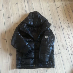 Svart pufferjacka med huva - Snygg svart pufferjacka med glansig finish, huva och dragkedja framtill. Jackan har en ficka med lock och Moncler-logga på armen. Perfekt för kalla dagar och ger en cool streetstyle-vibe.