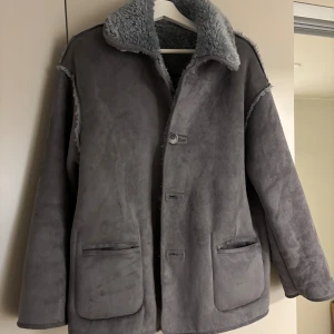 Grå teddyjacka med oversized passform - Säljer en grå teddyjacka med mjukt foder och stora fickor framtill. Jackan har en oversized passform, synliga sömmar och stängs med knappar. Perfekt för kalla dagar när du vill vara både varm och snygg.