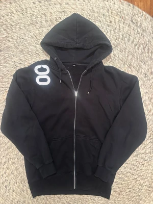 Svart hoodie från serien Johan falk med tryck och dragkedja - Använt i tv serien Johan falk på karaktären Jack Rollander. Svart hoodie med huva och coolt vitt tryck på ryggen som liknar en stiliserad insekt eller vingar. Framsidan har OG-logga i vitt på axeln och en hel dragkedja. Mjukt material och ribbade muddar, perfekt för chill dagar.