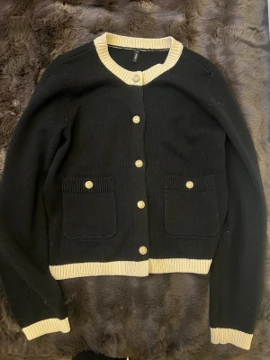 Svart Fin stickad cardigan med gult knappar  - Svart fin stickad cardigan med ljus beige rand längst halsen, armarna och längst ner på tröjan, tröjan har guldknappar med ett fint mönster på, tröjan är andvänt ett fåtal gånger