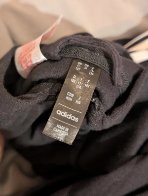 Svart Adidas hoodie med vita ränder - Svart hoodie från Adidas med klassiska vita ränder längs ärmarna och bröstet. Tröjan har huva, hel dragkedja framtill och långa ärmar med muddar. Perfekt för dig som gillar sportig och clean stil.  Materialet är mjukt och känns skönt mot huden. Pris kan diskuteras.