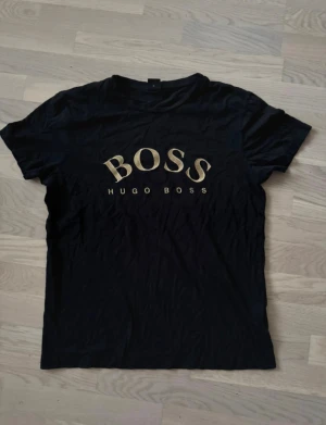 Svart Hugo Boss t-shirt med guldtryck - Svart t-shirt från Hugo Boss med stort BOSS-tryck i guld på bröstet. Klassisk rund hals och korta ärmar. Materialet är mjuk bomull som känns skönt mot huden. Perfekt för dig som gillar stilrena och lyxiga plagg med tydlig logga.