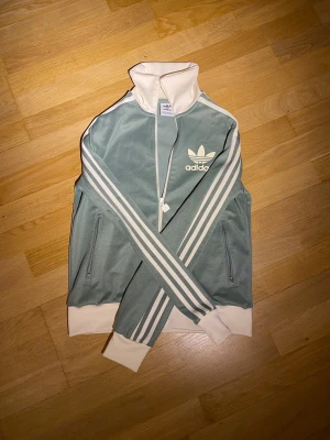 Adidas grönvit track jacket - Snygg Adidas track jacket i ljusgrönt med vita detaljer och klassiska tre ränder längs ärmarna. Jackan har hög krage, dragkedja framtill och två fickor med dragkedja. Adidas logga på bröstet och ribbade muddar i vitt. Knappt använd och i utmärkt skick 😀