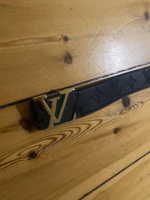 svart läderbälte - Svart läderbälte med ikoniskt mönster längs hela bandet och en stor, guldig LV-spänne framtill. Bältet är tillverkat i Spanien och har exklusiv känsla med tydliga detaljer och präglad logga på insidan.