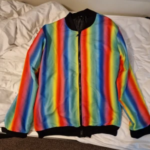 Färgglad randig bomberjacka - Säljer en riktigt färgstark bomberjacka med regnbågsrandigt mönster i rött, blått, grönt, gult och orange. Jackan har svarta muddar, krage och dragkedja framtill. Perfekt statement-plagg för dig som vill sticka ut i mängden. Den är handsydd och unik. Storlek stor L.