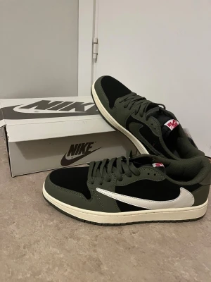 Nike Jordan 1 Low Olive/Black - Nike Jordan 1 Low sneakers i olivgrönt och svart med vit swoosh. Låg modell med snörning och platt sula. Materialmix av skinn och syntet, med detaljer i rött på tungan och insidan. Snygg och stilren färgkombo, perfekt för dig som gillar streetwear.