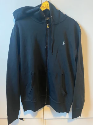 Svart hoodie från Polo Ralph Lauren - Svart hoodie från Polo Ralph Lauren med dragkedja och huva. Tröjan har två fickor framtill, snörning vid huvan och den klassiska Polo-loggan broderad på bröstet. Tillverkad i mjuk bomull, perfekt för chill dagar. Passformen är normal och ärmarna är långa.