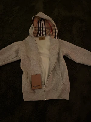 Grå hoodie från Burberry med dragkedja - Snygg grå hoodie från Burberry helt ny och oanvänd med klassiskt rutmönstrat foder i huvan. Dragkedja framtill, två fickor och ribbade muddar. Perfekt för dig som gillar stilrena plagg med exklusiv touch. Materialet känns mjukt och bekvämt. Priset är ej hugget i sten 