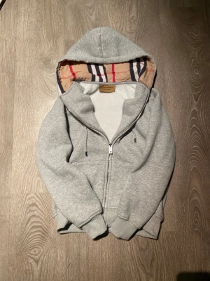 Grå hoodiejacka från Burberry - Snygg grå hoodiejacka från Burberry med dragkedja och klassisk rutmönstrad insida i huvan. Jackan har snörning vid huvan och ribbade muddar. Perfekt för dig som gillar stilren design med en touch av lyx.