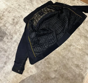 Svart bomberjacka från Hugo Boss - Snygg svart bomberjacka från Hugo Boss med quiltat foder och rutigt innerfoder i grått och gult. Jackan har dragkedja, knappar vid ärmslut och detaljer i gult längs insidan. Perfekt för dig som gillar stilrena och klassiska jackor med modern touch.