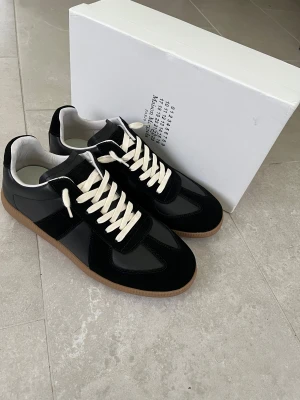 Maison Margiela svarta sneakers i skinn - Maison Margiela sneakers i svart skinn och mocka med vita snören och klassisk gummisula. Snygg minimalistisk design med diskreta detaljer och ikonisk logotyp på sulan. Perfekta för dig som gillar stilrena och exklusiva sneakers.