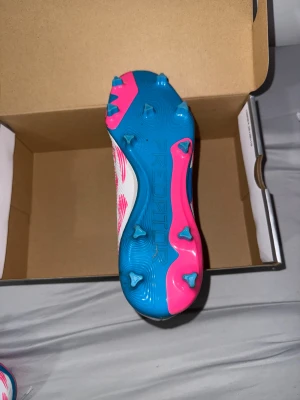 Adidas Predator fotbollsskor vit/blå/rosa - Säljer ett par Adidas Predator fotbollsskor med snyggt mönster i vitt, blått och rosa. Skorna har snörning, platt sula med dobbar och är tillverkade i syntetmaterial. Perfekt för dig som vill sticka ut på planen med en modern och sportig look.