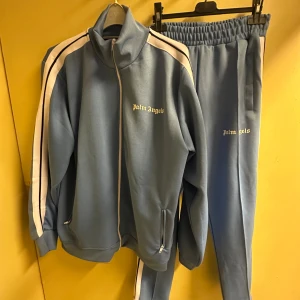  Palm Angels tracksuit, blue. Size:,XL - Säljer en blå tracksuit från Palm Angels med vita ränder längs ärmarna och ben. Tröjan har hög krage, lång ärm, dragkedja framtill och  fickor med zip. Matchande byxor i Palm Angelsk klassiska utförande.. Ikonisk Palm Angels-logga på bröstet och lår. Perfekt för dig som gillar streetwear och sportig stil. 