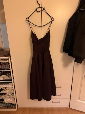 Mörkbrun långklänning med smala axelband - Säljer en elegant mörkbrun långklänning med smala axelband och omlott-detalj upptill. Klänningen har en v-ringad överdel och ett utsvängt, veckat kjolparti som ger en snygg siluett. Perfekt för dig som vill sticka ut med en stilren look.