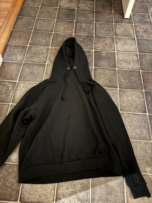 Svart hoodie från 157 med huva - Svart hoodie från 157 i storlek L sitter som M. Klassisk modell med huva och snörning, tillverkad i mjuk bomull. Perfekt för chill dagar och enkel att matcha med jeans eller joggers. Snygg och enkel design utan tryck eller mönster.