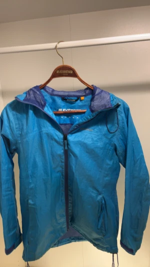 Blå vindjacka från Everest, strl 36 - Snygg blå vindjacka från Everest med huva och meshfoder. Jackan har dragkedja framtill, två fickor med dragkedja och justerbar nederkant med snörning. Perfekt för blåsiga dagar och outdoor-aktiviteter.