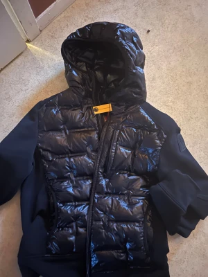 Svart Parajumpers Techno Hybrid jacka M - Säljer en svart Parajumpers Techno Hybrid jacka i storlek M. Jackan har glansig quiltad framsida och huva, med släta svarta ärmar och sidor. Dragkedja framtill och ikonisk gul detalj vid halsen. Perfekt för dig som vill ha en snygg och sportig look.