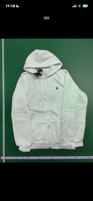 Vit hoodie från Polo Ralph Lauren - Vit hoodie från Polo Ralph Lauren med dragkedja och huva med snörning. Klassisk design med broderad logga på bröstet och två fickor framtill. Mjuk och skön bomullsmix, perfekt för chill dagar. Ribbstickade muddar vid ärmslut och nederkant.