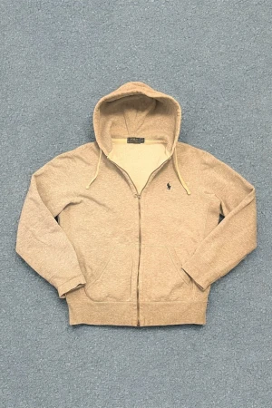 Grå hoodie från Polo Ralph Lauren - Snygg beige hoodie från Polo Ralph Lauren med dragkedja, huva med snören och mjukt fleecefoder. Klassisk logga broderad på bröstet och två stora fickor framtill. Perfekt för dig som gillar stilren och bekväm streetwear.