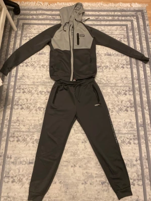 Grå tracksuit från mckenzie - Snygg tvåfärgad hoodie från Mckenzie i mörkgrått och ljusgrått. Hoodien har dragkedja, fickor med dragkedja och en justerbar huva med snören. Materialet känns mjukt och sportigt, perfekt för chill eller träning. Märkeslogga på bröstet och ärmen.
