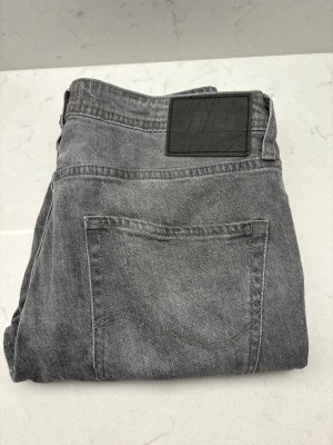 Grå jeans från Jack & Jones - Snygga grå jeans från Jack & Jones med klassisk femficksdesign och svart patch bak i midjan. Jeansen har en rak passform och är tillverkade i slitstarkt bomullsmaterial. Perfekt för dig som gillar en enkel och stilren look.