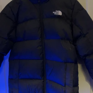 Svart pufferjacka från The North Face - Säljer en svart pufferjacka från The North Face med hög krage och diskret logga på bröstet. Jackan har lång ärm, dragkedja framtill och är riktigt fluffig med breda paneler. Perfekt för kalla dagar och har en clean, urban vibe.