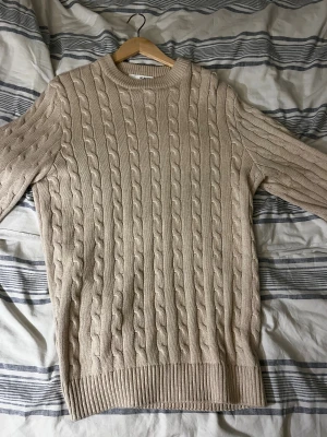 Beige kabelstickad tröja Selected Homme - Kabelstickad beige tröja från Selected Homme med rund halsringning och ribbade muddar. Tröjan har en klassisk, chunky look och är perfekt för lager-på-lager under kyliga dagar. Passar dig som gillar stilren och enkel design. Tröjan är endast använd en gång. 