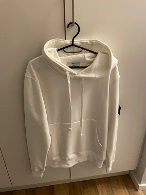 Vit hoodie från Stone Island - Vit hoodie från Stone Island med klassisk logopatch på vänster ärm. Tröjan har huva med snörning, stor magficka och ribbade muddar. Mjuk bomullskvalitet och relaxed passform, perfekt för chill dagar. Snygg och enkel design som funkar till allt.Helt nytt tags medföljer 