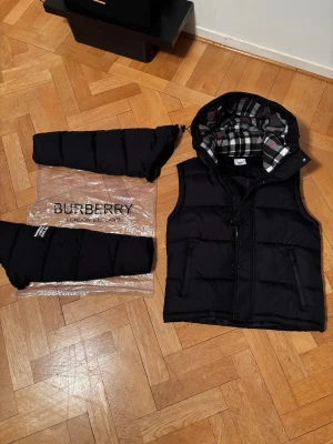Svart pufferjacka från Burberry - Svart pufferjacka från Burberry med avtagbara ärmar så du kan bära den som väst. Jackan har huva med klassiskt rutigt foder i grått, vitt och rött, samt två dragkedjefickor fram. Burberry-logga på vänster ärm. Perfekt för kalla dagar. 