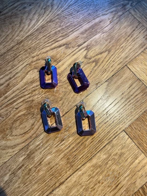 Chunky örhängen i lila och rött - Tre par statement örhängen i chunky stil, ett par i metallic-lila och ett par i metallic-rött. Båda har geometrisk, rektangulär form och guldfärgade detaljer. Materialet är plast med glansig finish och örhängena har pluppar i transparent silikon.