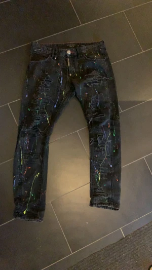 Philipp Plein svarta jeans med färgstänk - Unika svarta jeans från Philipp Plein med färgstänk i neonfärger och slitna detaljer framtill. Slim fit-modell med logotyp på bakfickan och coolt tryck på insidan av midjan. Perfekt för dig som vill sticka ut med din stil.