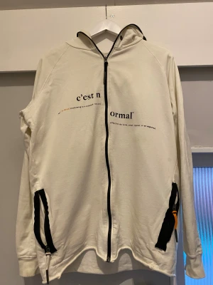 Vit hoodie från C'est Normal - Vit hoodie från Jon Olssons egna ”C'est Norma”  med svart tryck på bröstet och coola svarta detaljer vid fickorna. Tröjan har hel dragkedja, huva och ribbade muddar. Tillverkad i 100% bomull, med orange etikett på ärmen och textdetaljer framtill. En limiterad kollektion som inte längre finns kvar, nypris ca 1200kr (Pris går att diskutera vid smidig affär)