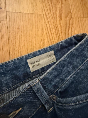 High rise relaxed jeans från Vero Moda - Blå jeans med hög midja och avslappnad passform från Vero Moda. Modellen har raka, breda ben och klassiska fickor både fram och bak. Jeansen är tillverkade i ett mjukt denimtyg och har bälteshällor samt knapp och dragkedja framtill.