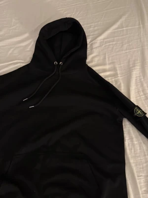 Svart hoodie från Stone Island - Svart hoodie från Stone Island med klassisk patch på vänster ärm. Hoodien har dragsko i huvan och en stor magficka. Tillverkad i mjukt bomullstyg och har en clean, stilren look som passar perfekt till streetwear. Ikonisk logga på ärmen och etikett i nacken.