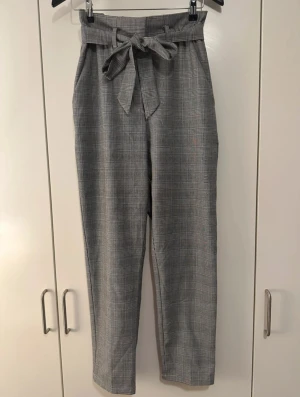 Grå rutiga kostymbyxor från H&M - Snygga grå kostymbyxor från H&M i storlek 36 med klassiskt rutigt mönster. Byxorna har hög midja, knytband i midjan och raka ben. Perfekt för dig som gillar en stilren och trendig look. Materialet är mjukt och bekvämt, och byxorna har praktiska fickor.