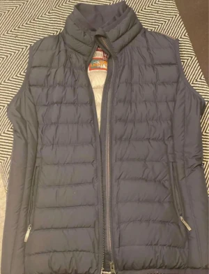 Mörkblå dunväst  - Säljer en mörkblå dunväst från med quiltad design och hög krage. Västen har två sidofickor med dragkedja och en smidig dragkedja framtill. Perfekt för lager-på-lager och chill stil.