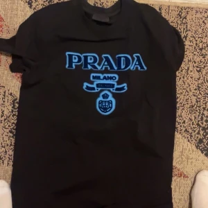 Prada tröja  - Svart t-shirt från Prada med stort blått PRADA-tryck och Milano-emblem på bröstet. Klassisk rund hals och korta ärmar. Snygg och stilren design med kontrasterande färger som gör att loggan verkligen poppar.
