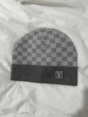 Grå rutig halsduk från Louis Vuitton - Snygg halsduk från Louis Vuitton i grått och vitt med klassiskt rutigt mönster. Halsduken har en rektangulär form och är dekorerad med LV-logga i ena hörnet. Perfekt för dig som vill ha en stilren accessoar med lyxig känsla.