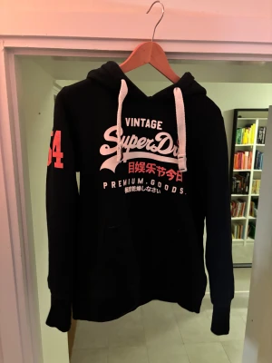 Marinblå Superdry hoodie med tryck - Marinblå hoodie från Superdry med stort vitt och rött tryck på bröstet och siffran 54 i rött på ärmen. Tröjan har huva med vita snören och en klassisk känguruficka framtill. Insidan är mjukt fodrad för extra komfort.