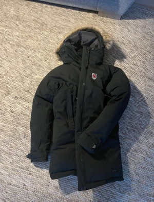 Svart dunjacka med päls från Fjällräven - Säljer en svart dunjacka från Fjällräven med snygg päls på huvan och klassisk röd logga på bröstet. Jackan har dragkedja, knappar och flera fickor. Perfekt för kalla vinterdagar och har en stilren, längre passform.                 Den är i storlek M, men den som har på sig jackan är 167. Jackan skulle passa S även! Personen som har på sig jackan har vanligtvis S. Det är dam storlek!! #fjällräven #jacka #fjällrävenjacka #sellout 