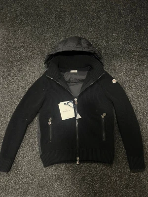 Svart Moncler pufferjacka med huva - Svart pufferjacka från Moncler med stickade ärmar och quiltad kropp. Jackan har en stor huva, två dragkedjeförsedda fickor och ribbade muddar. Materialmix av dun och stickad polyester ger både värme och stil. Perfekt för dig som vill ha en snygg och funktionell jacka😊