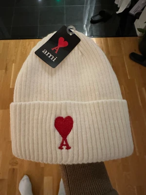 Vit ribbad mössa från Ami - Snygg vit ribbstickad mössa från Ami med röd broderad logga i form av ett hjärta och bokstaven A framtill. Mössan har uppvikt kant och är tillverkad i mjukt material som håller dig varm och stilren under kalla dagar. Perfekt nu när kallare tider kommer. Såklart äkta. Priset är inte hugget i sten!