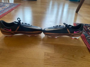 Nike Phantom GT fotbollsskor svart/koppar - Säljer ett par Nike Phantom GT fotbollsskor i svart och koppar med vita och rosa detaljer. Skorna har snörning, platt sula med fasta dobbar och en sportig design. Ovandelen är i syntetmaterial och skon har en rund tå. Perfekta för dig som vill sticka ut på planen. Är även nästan oanvänd bara använda enstaka gånger