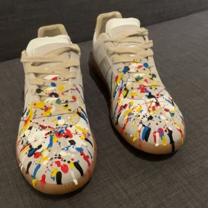 Maison margela gats  - Unika vita sneakers från Maison margela med färgstänk i rött, blått, gult och svart. Skorna har beige snörning, gummisula och är tillverkade i skinn. Perfekt för dig som vill sticka ut med en kreativ och cool stil.