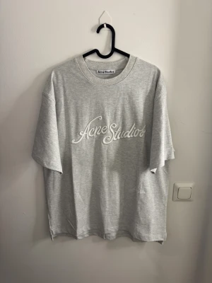 Grå t-shirt Acne Studios broderad logga unisex S - Snygg grå t-shirt från Acne Studios med broderad logga framtill i vitt. Modellen är loose fit med korta ärmar och har en liten slits i nederkanten. Tillverkad i mjuk bomull som känns skön mot huden. Perfekt för dig som gillar stilrena och trendiga plagg.