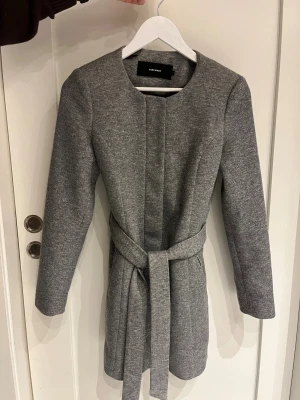 Grå kappa med bälte från Vero Moda - Halvlång grå kappa från Vero Moda i stilren design med rund hals och bälte i midjan. Jackan har dold dragkedja och knappar framtill. Materialet är mjukt med en blandning av polyester 85% och bomull 15%. Fint skick.
